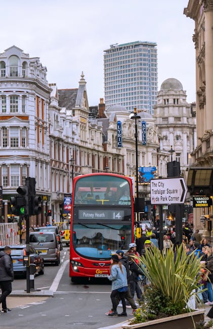 A red double-decker bus parked on a busy street in central Putney, London, with pedestrians crossing in front of it and shoppers walking along the sidewalk near various retail stores and cafes. The bu