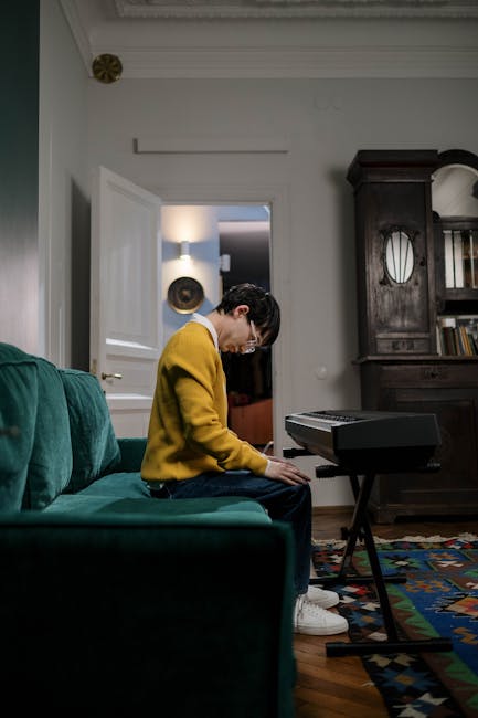 The image shows a black upright piano positioned inside a spacious room with large rectangular windows on the left side, allowing natural light to illuminate the space. The piano is placed on a tiled