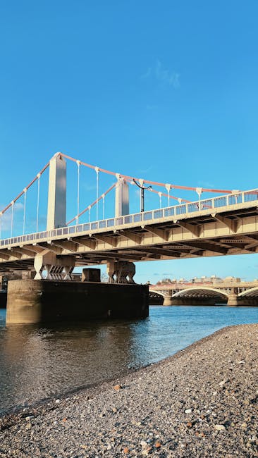 The image depicts a section of a suspension bridge spanning over a river under a clear blue sky, with a visible support structure and cables. In the foreground, a gravelly riverbank is present, and a portion of a moving van or truck is partially visible on the right side of the image, parked close to the bridge's base. The scene is set during daylight, with natural lighting highlighting the bridge's metal framework and the surrounding environment. This setting is relevant to home relocation and furniture transport, suggesting a context where loading or unloading of moving boxes and household items could occur near the bridge area, with Removal Company Putney potentially utilizing such Thames-side access routes for efficient house removals and logistics.