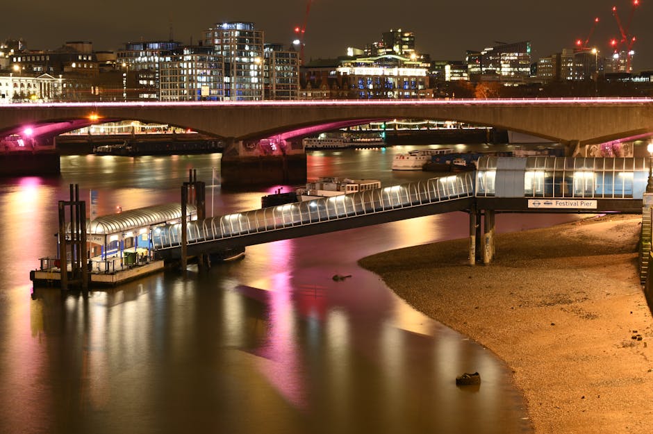 A nighttime view of the Thames river in Putney with illuminated bridges and modern city buildings in the background. In the foreground, a glass-enclosed pier taxi shelter extends over the water, supported by concrete pillars, with a small dock beneath. Next to the shelter, a moving trolley loaded with cardboard boxes covered in plastic wrap is being transported, possibly as part of a home relocation service. Several larger cardboard boxes and plastic-wrapped packages are positioned nearby on the riverbank, indicating packing and moving activities typical of furniture transport and packing and moving operations. The scene captures the loading process along the river during evening hours, with reflections of city lights shimmering on the calm water surface, and the environment lit by ambient street and bridge lighting, framing the ongoing logistics involved in house removals facilitated by Removal Company Putney near the Thames. The photo emphasizes the connection between waterfront access and efficient moving logistics for residential relocations in the area.