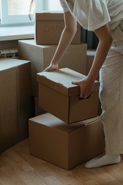 Two movers wearing dark uniforms and protective glasses are engaged in the loading process of a home relocation on a paved street. They are carefully positioning a large, rectangular, green upholstere