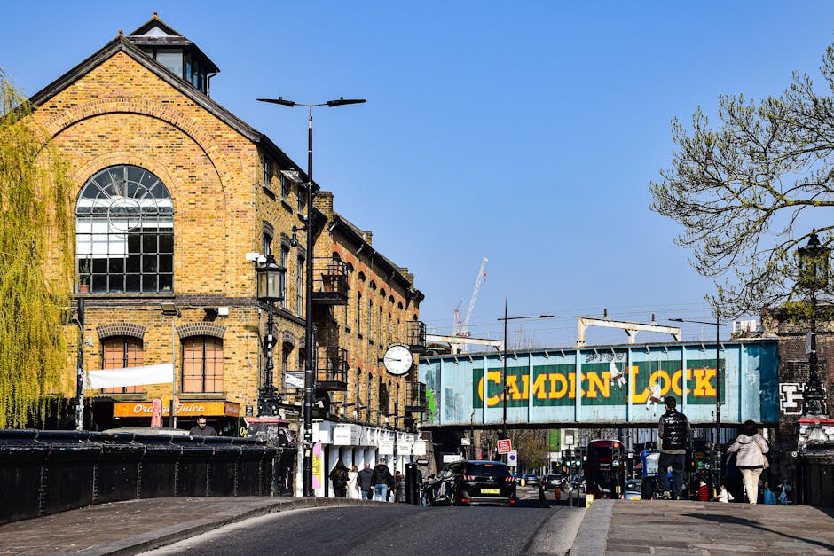 A modern passenger train with a sleek yellow and grey exterior is shown traveling along railway tracks beneath an old brick railway bridge. The train is moving through a green, tree-lined area with de